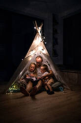 Vater mit kleinen Kindern sitzt abends im Zelt im Schlafzimmer und liest ein Buch. - HPIF21363