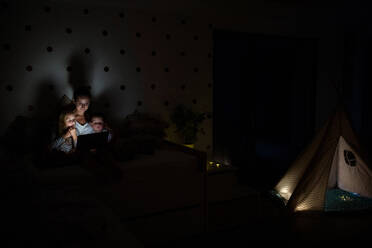 Junge Mutter mit Kindern, die abends im Schlafzimmer sitzen und einen Laptop benutzen. - HPIF21369