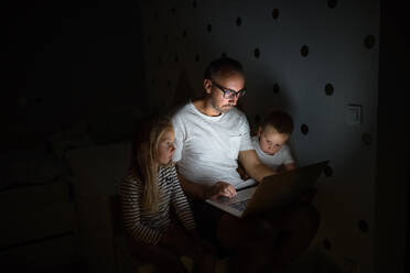 Father with small children sitting indoors in bedroom in the evening, using laptop. - HPIF21372