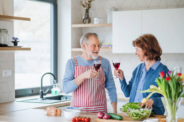 A portrait of senior couple in love indoors at home, clinking glasses with wine when cooking. - HPIF21457