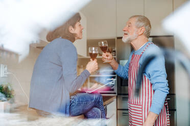 A portrait of happy senior couple in love indoors at home, clinking glasses. Shot through glass. - HPIF21468