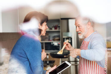 A portrait of happy senior couple in love indoors at home, cooking. Shot through glass. - HPIF21469