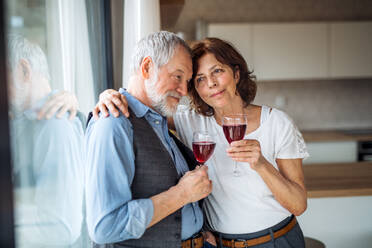 A senior couple with wine standing indoors at home, standing by window. - HPIF21479