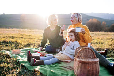 Glückliches kleines Mädchen mit Mutter und Großmutter beim Picknick in der Natur bei Sonnenuntergang. - HPIF21656