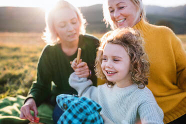 Glückliches kleines Mädchen mit Mutter und Großmutter beim Picknick in der Natur bei Sonnenuntergang. - HPIF21659