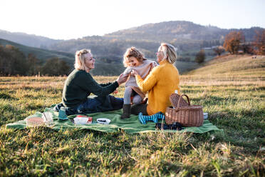 Glückliches kleines Mädchen mit Mutter und Großmutter beim Picknick in der Natur bei Sonnenuntergang. - HPIF21660