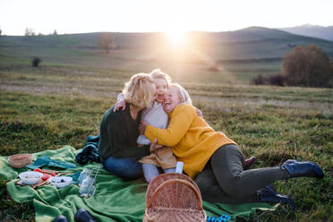 Glückliches kleines Mädchen mit Mutter und Großmutter beim Picknick in der Natur bei Sonnenuntergang, die Spaß haben. - HPIF21663