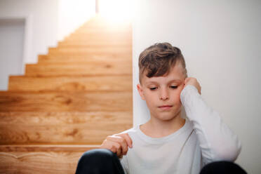 A sad small unhappy boy sitting on the floor by the stairs. Copy space. - HPIF21717