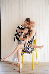 Young woman with two children in black and white clothes sitting on yellow chair against white wooden wall. - HPIF21726