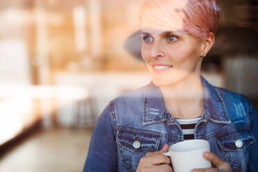 A close-up portrait of young attractive woman with coffee standing by window at home. Shot through glass. - HPIF21729