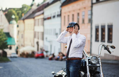 Ein fröhlicher älterer Geschäftsmann mit Motorrad in der Stadt, der seinen Helm aufsetzt. - HPIF21755