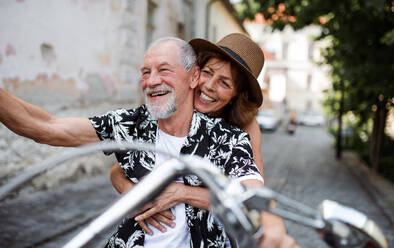 A front view of cheerful senior couple travellers with motorbike in town. - HPIF21764