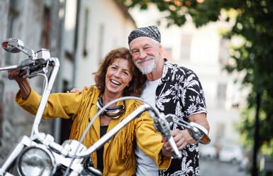 A front view of cheerful senior couple travellers with motorbike in town. - HPIF21803