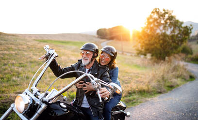 A cheerful senior couple travellers with motorbike in countryside at sunset. - HPIF21827