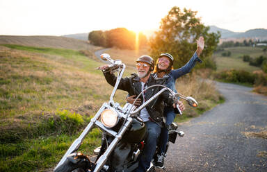 A cheerful senior couple travellers with motorbike in countryside at sunset. - HPIF21831