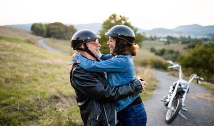 A front view portrait of cheerful senior couple travellers with motorbike in countryside, hugging. - HPIF21836