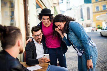 Eine Gruppe junger, fröhlicher Freunde mit Smartphone sitzt in einem Straßencafé und unterhält sich. - HPIF21990