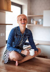 A young attractive woman with a coffee sitting on a counter in a kitchen at home, laughing. - HPIF22095
