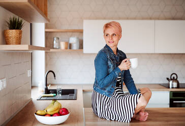 A young attractive woman with a coffee sitting on a counter in a kitchen at home. - HPIF22096