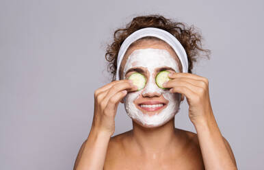 Ein Porträt einer jungen Frau mit Gesichtsmaske und Gurke in einem Studio, Schönheit und Hautpflege-Konzept. - HPIF22110