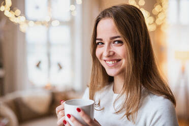 Porträt einer glücklichen jungen Frau, die zu Weihnachten zu Hause ist und eine Tasse Kaffee in der Hand hält. - HPIF22220