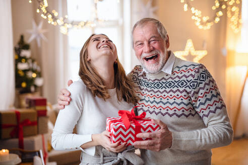 Lachender älterer Mann mit junger Frau zu Hause an Weihnachten, mit Geschenk in der Hand. - HPIF22233