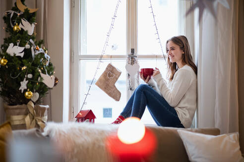 Porträt einer jungen Frau zu Hause an Weihnachten, die mit einem Tee auf der Fensterbank sitzt. - HPIF22270