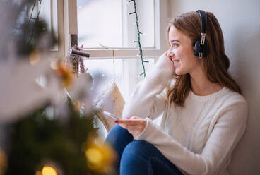 Portrait of young woman sitting indoors at home at Christmas, using smartphone and headphones. - HPIF22271
