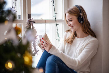 Porträt einer jungen Frau, die zu Weihnachten zu Hause sitzt und ein Smartphone und Kopfhörer benutzt. - HPIF22272