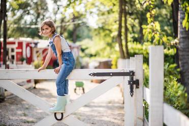 Ein Porträt eines glücklichen kleinen Mädchens auf dem Bauernhof der Familie, das auf einem Holztor sitzt. - HPIF22294