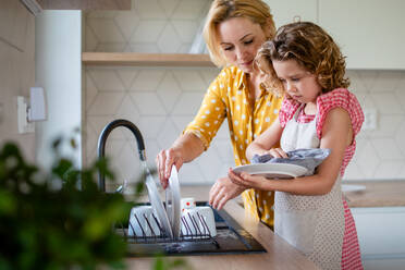 A cute small girl with mother indoors in kitchen at home, washing up the dishes. - HPIF22355