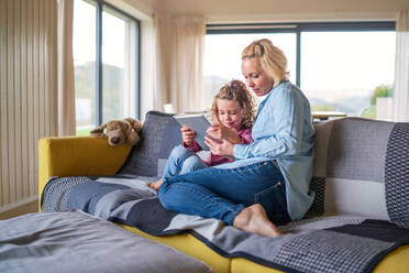 A front view of cute small girl with mother on sofa indoors at home, using tablet. - HPIF22359