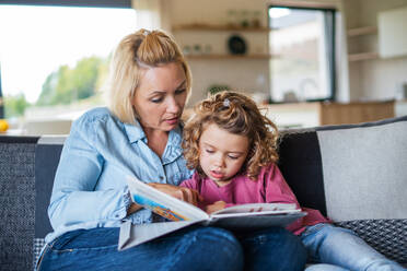 A cute small girl with mother sitting on sofa indoors at home, reading story book. - HPIF22368