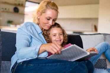 A cute small girl with mother sitting on sofa indoors at home, reading story book. - HPIF22370