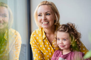 A cute small girl with mother indoors at home, looking out through window. - HPIF22412