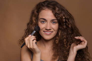 Porträt einer jungen Frau mit Schminkpinsel in einem Studio vor braunem Hintergrund. - HPIF22514