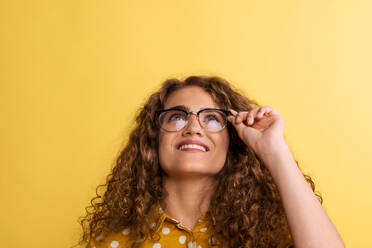 Porträt einer jungen glücklichen Frau mit Brille in einem Atelier auf gelbem Hintergrund, die nach oben schaut. - HPIF22526