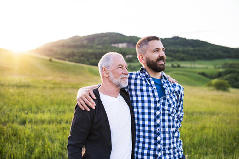 Ein Porträt eines lachenden erwachsenen Hipster-Sohns mit seinem älteren Vater in der Natur bei Sonnenuntergang, die Arme umeinander gelegt. - HPIF22551