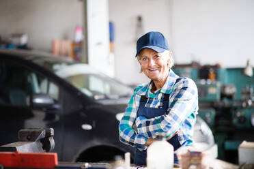 Eine ältere Frau steht mit verschränkten Armen in einer Werkstatt und repariert ein Auto. - HPIF22734