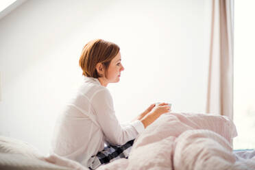 Eine junge Frau mit Nachthemd sitzt morgens in einem Schlafzimmer auf dem Bett und hält eine Tasse. - HPIF22809