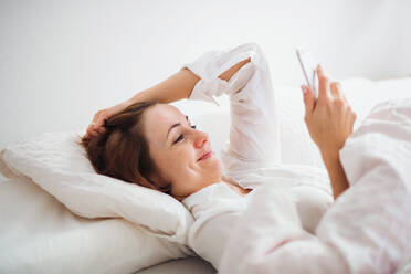 A happy young woman lying in bed indoors in the morning in a bedroom, using smartphone. - HPIF22811