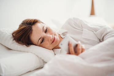 A happy young woman lying in bed indoors in the morning in a bedroom, using smartphone. - HPIF22812