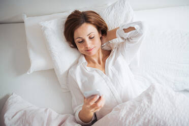 A top view of happy young woman lying in bed indoors in the morning in a bedroom, using smartphone. - HPIF22813