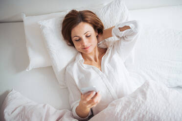 Eine Draufsicht auf eine glückliche junge Frau, die morgens in einem Schlafzimmer im Bett liegt und ein Smartphone benutzt. - HPIF22813