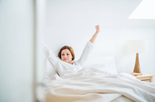 Eine glückliche junge Frau sitzt morgens in einem Schlafzimmer im Bett und streckt sich. - HPIF22816