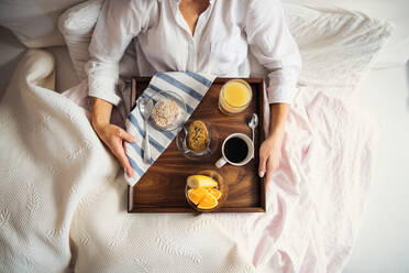 A midsection of woman lying in bed with coffee and breakfast indoors in the morning in a bedroom, using smartphone. A top view. - HPIF22818