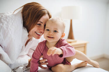 Eine glückliche junge Mutter mit ihrer kleinen Tochter, die am Morgen im Haus auf dem Bett sitzt und spielt. - HPIF22829