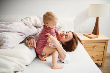 Eine glückliche junge Mutter mit ihrer kleinen Tochter, die am Morgen im Haus auf dem Bett sitzt und spielt. - HPIF22830