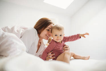 Eine glückliche junge Mutter mit ihrer kleinen Tochter, die am Morgen im Haus auf dem Bett sitzt und spielt. - HPIF22831
