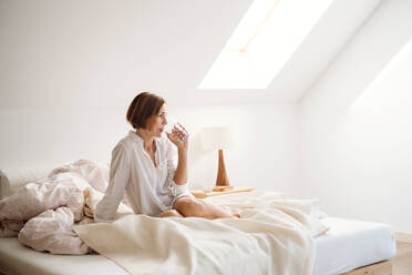 Eine junge Frau mit Nachthemd sitzt morgens im Schlafzimmer auf dem Bett und trinkt Wasser. - HPIF22843